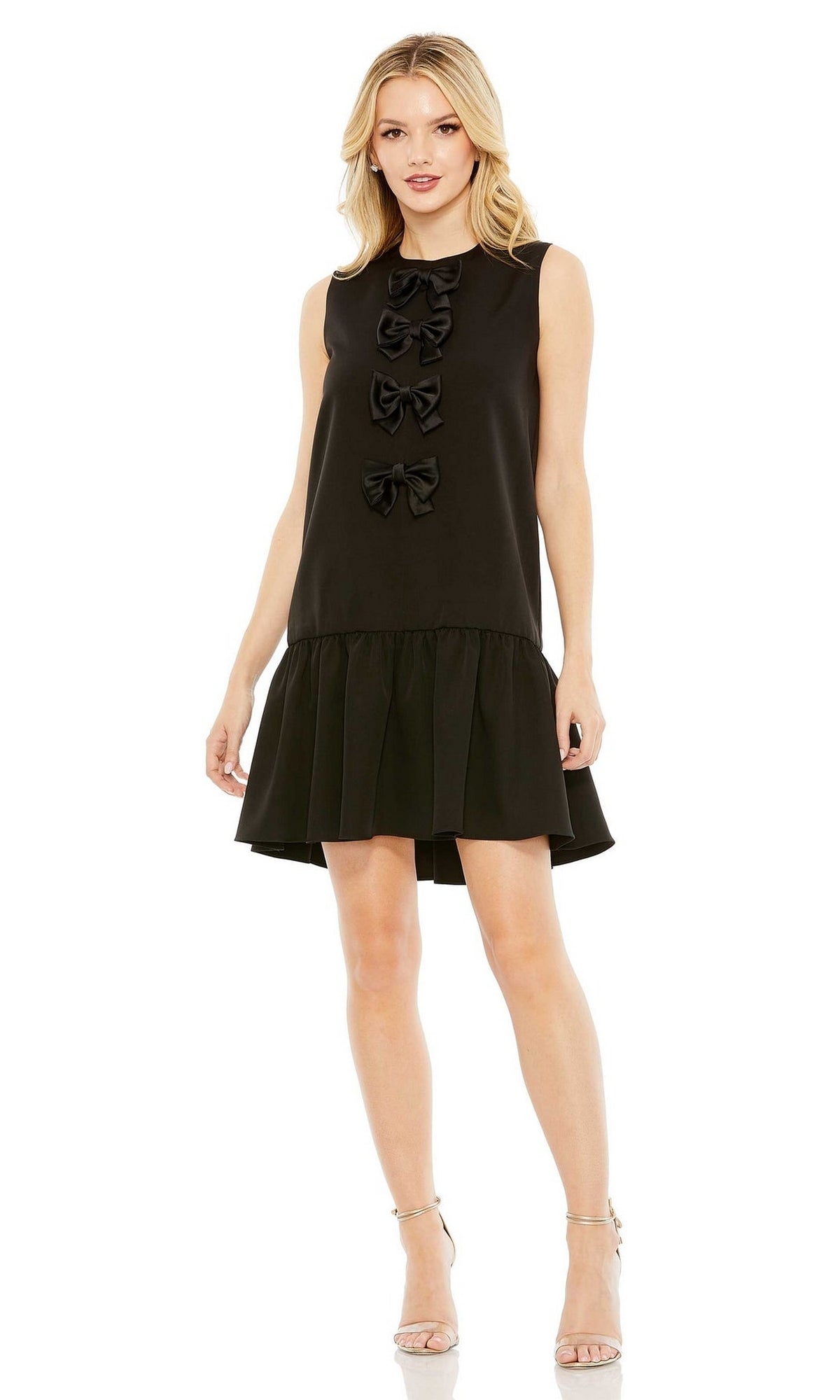 A woman stands against a white background wearing the Mac Duggal Short Wedding Guest Dress 27032—a sleeveless party dress with three front bows and a pleated hem—paired with strappy high heels.